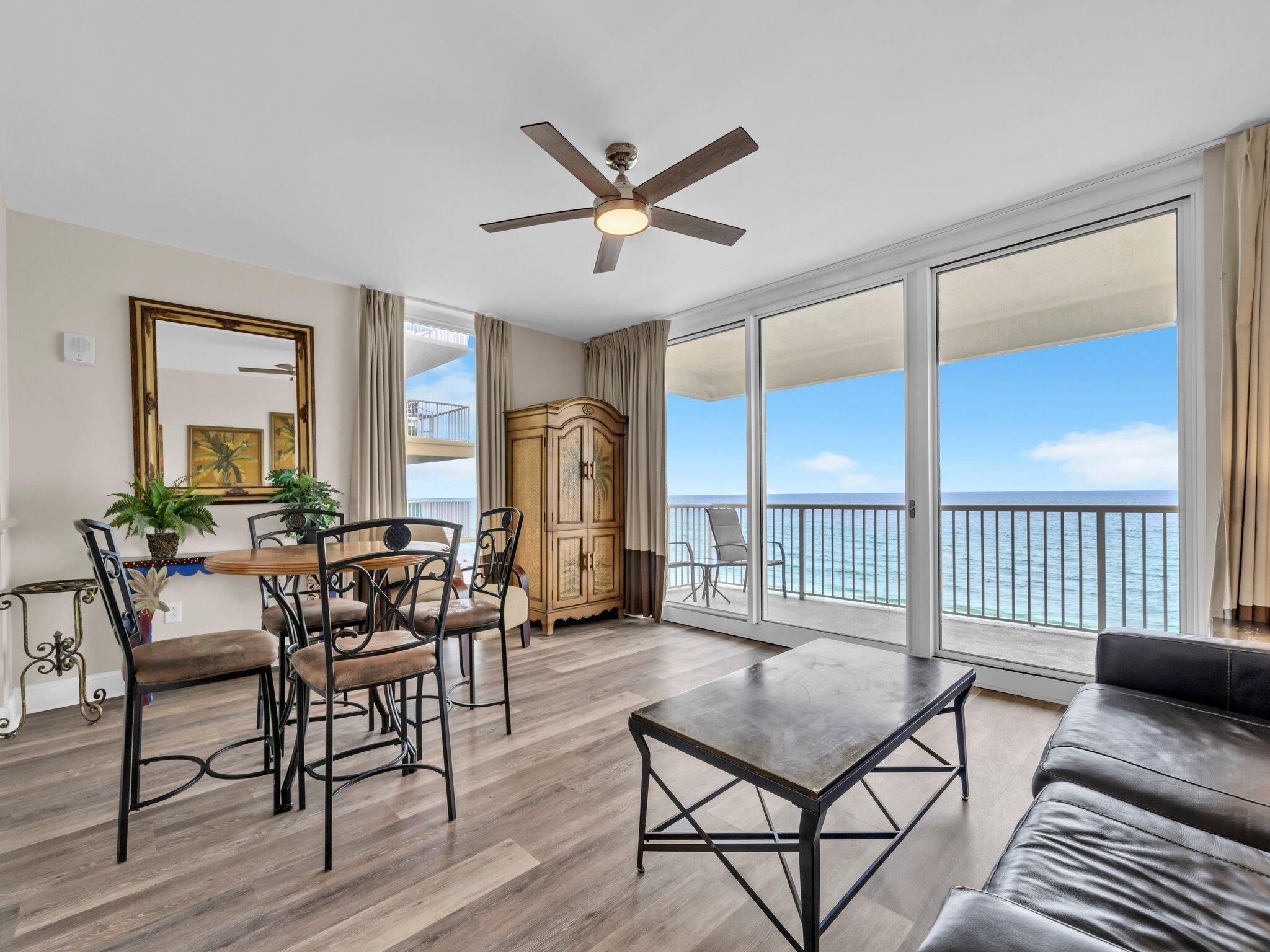 A spacious living room with a balcony overlooking the sea, a ceiling fan, and a dining area with chairs and a table.