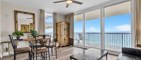 A spacious living room with a balcony overlooking the sea, a ceiling fan, and a dining area with chairs and a table.