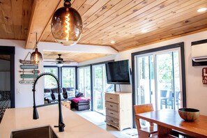 A modern kitchen with a wooden ceiling, a dining area with a wooden table and chairs, and a large window providing a view of the outdoors.