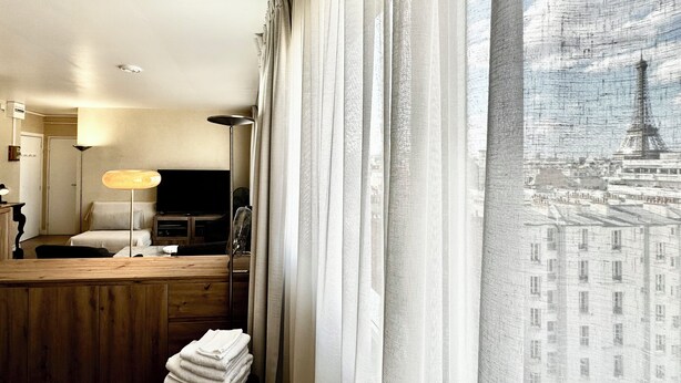 A hotel room with a bed, bedside table, lamp, and a view of a cityscape through sheer curtains. - Studio with beautiful view of the Eiffel Tower, 10 minutes' walk from the Champs-de-Mars (Paris)