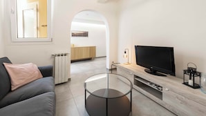 Ett modernt vardagsrum med en soffa, ett soffbord i glas, en tv på ett träställ och en välvd dörröppning som leder till ett annat rum.