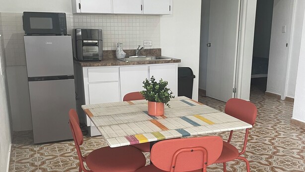 Een kleine keuken met witte kastjes, een magnetron en een elektrische oven. Er is een eettafel met vier rode stoelen en een potplant. De vloer heeft betegeling met een patroon.