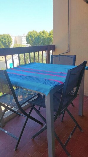 A balcony with a table and chairs, a blue tablecloth, and a view of trees and buildings.