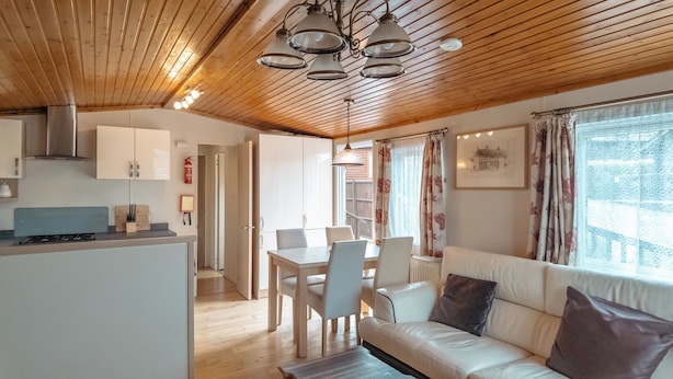 A cozy interior with wooden ceiling beams, a dining area with a table and chairs, a sofa with cushions, and a kitchen with cabinets and a stove.
