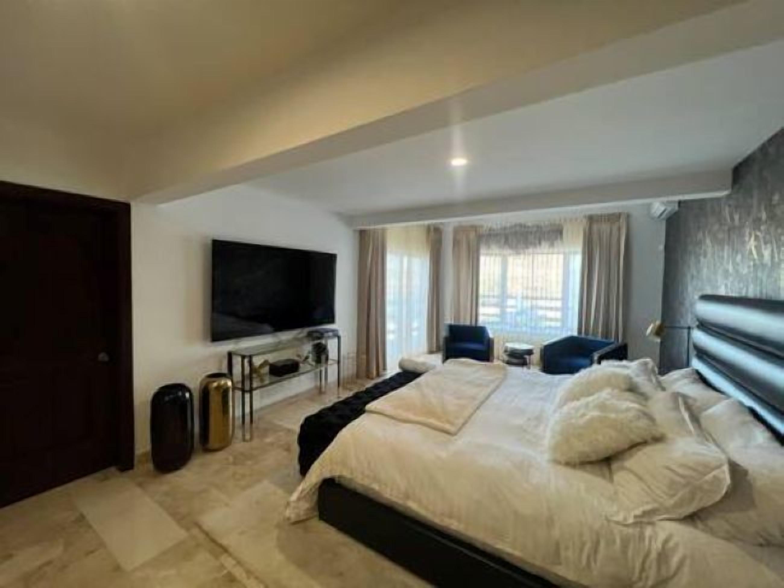 Ein modernes Schlafzimmer mit einem großen Bett, einem flachbildfernseher an der Wand und Blick ins Freie durch große Fenster.