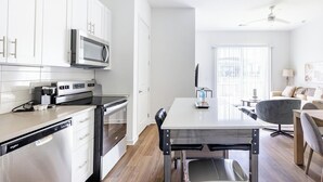 A modern kitchen with white cabinetry, a microwave, a stove, and a dishwasher. There is a dining table with chairs and a water bottle on it. The room has a ceiling fan and a window with blinds.