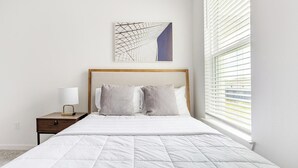 A neatly made bed with a white quilt, a wooden headboard, and two pillows. There is a nightstand with a lamp and a wall-mounted abstract art piece.