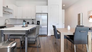 Una cocina moderna con armarios blancos, un refrigerador de acero inoxidable y un comedor con mesa de madera y sillas grises.