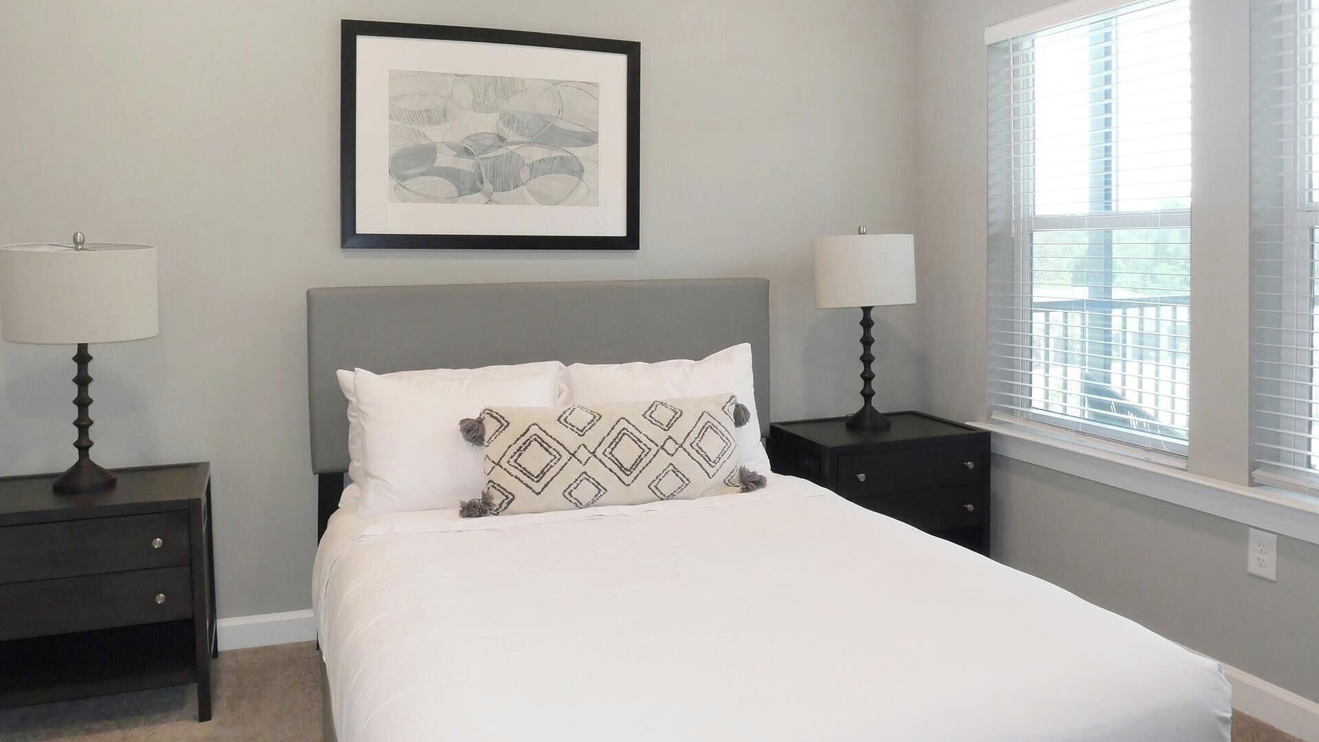 A bedroom with a bed, two nightstands, a framed picture on the wall, and a window with blinds.