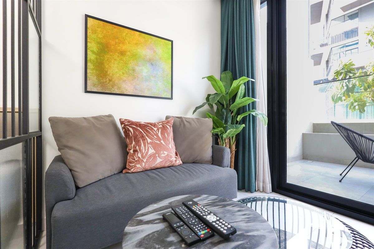 A modern living room with a grey sofa, a plant, and a large window with a view of buildings.