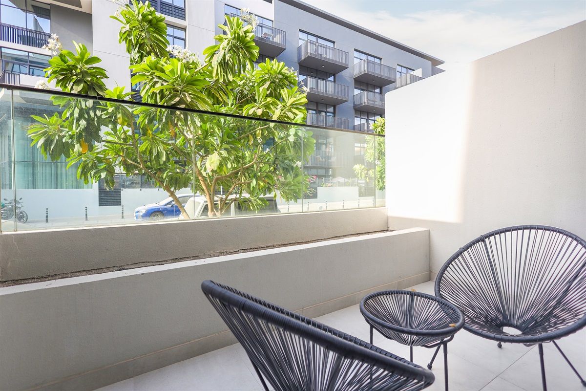 Een balkon met twee zwarte rieten stoelen, een glazen hekwerk en uitzicht op een gebouw met balkons en een boom.