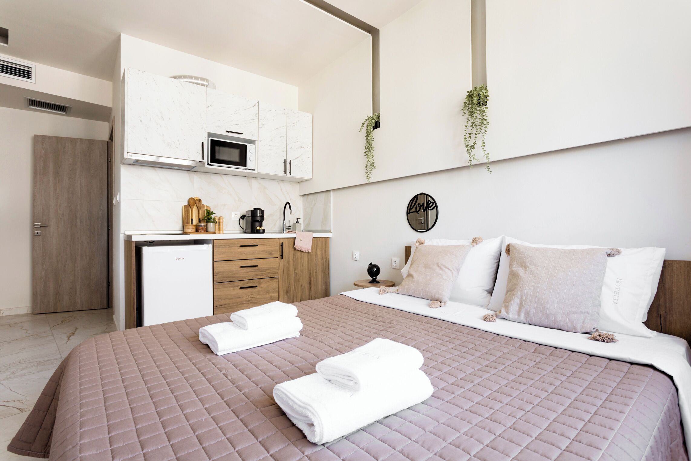 A modern hotel room with a bed, a small kitchenette, and a wall-mounted plant.