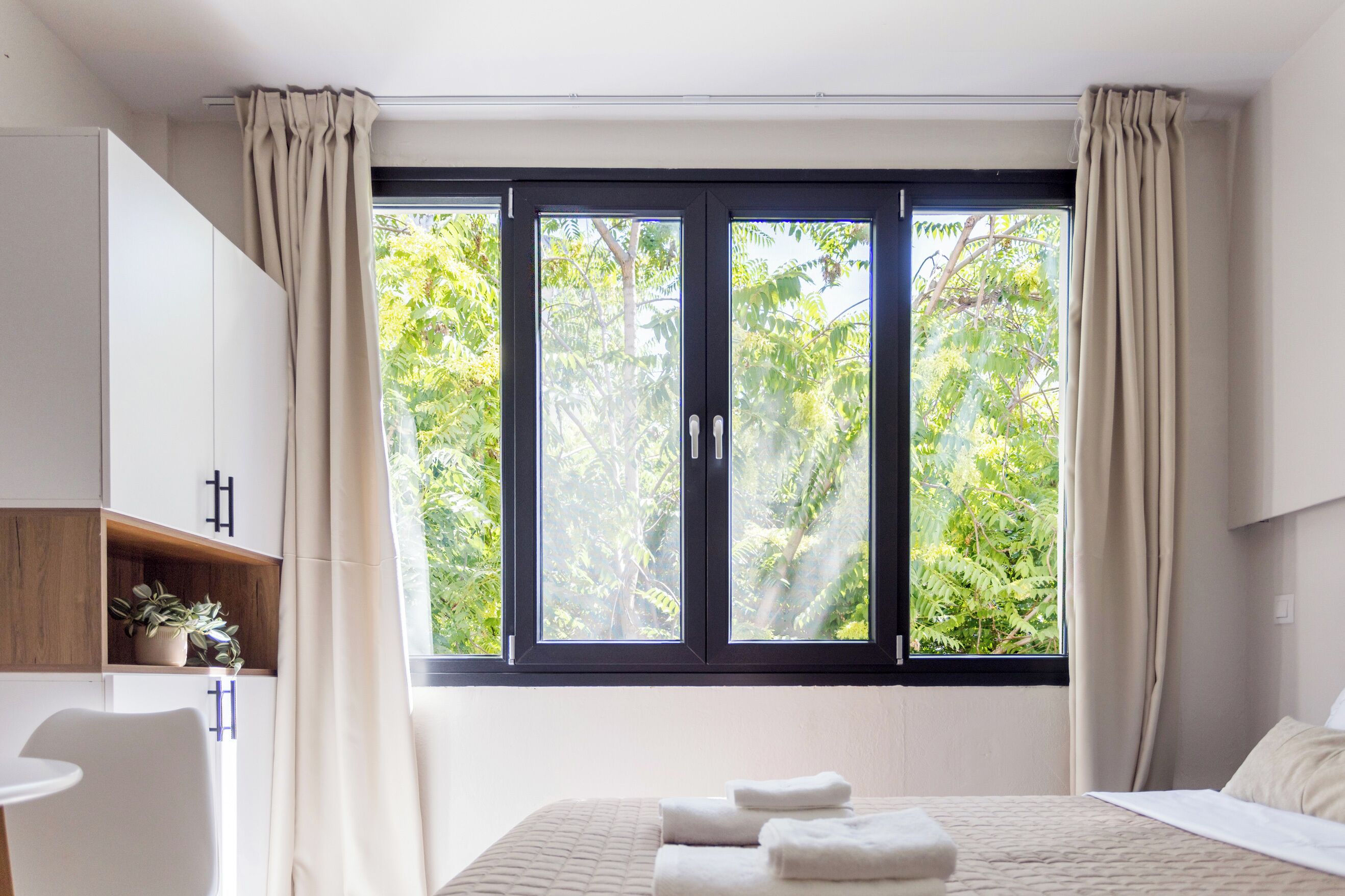 Ein Schlafzimmer mit einem Bett, zwei Nachttischen, einem Sessel, einem Fenster mit Vorhängen und Blick ins Grüne.