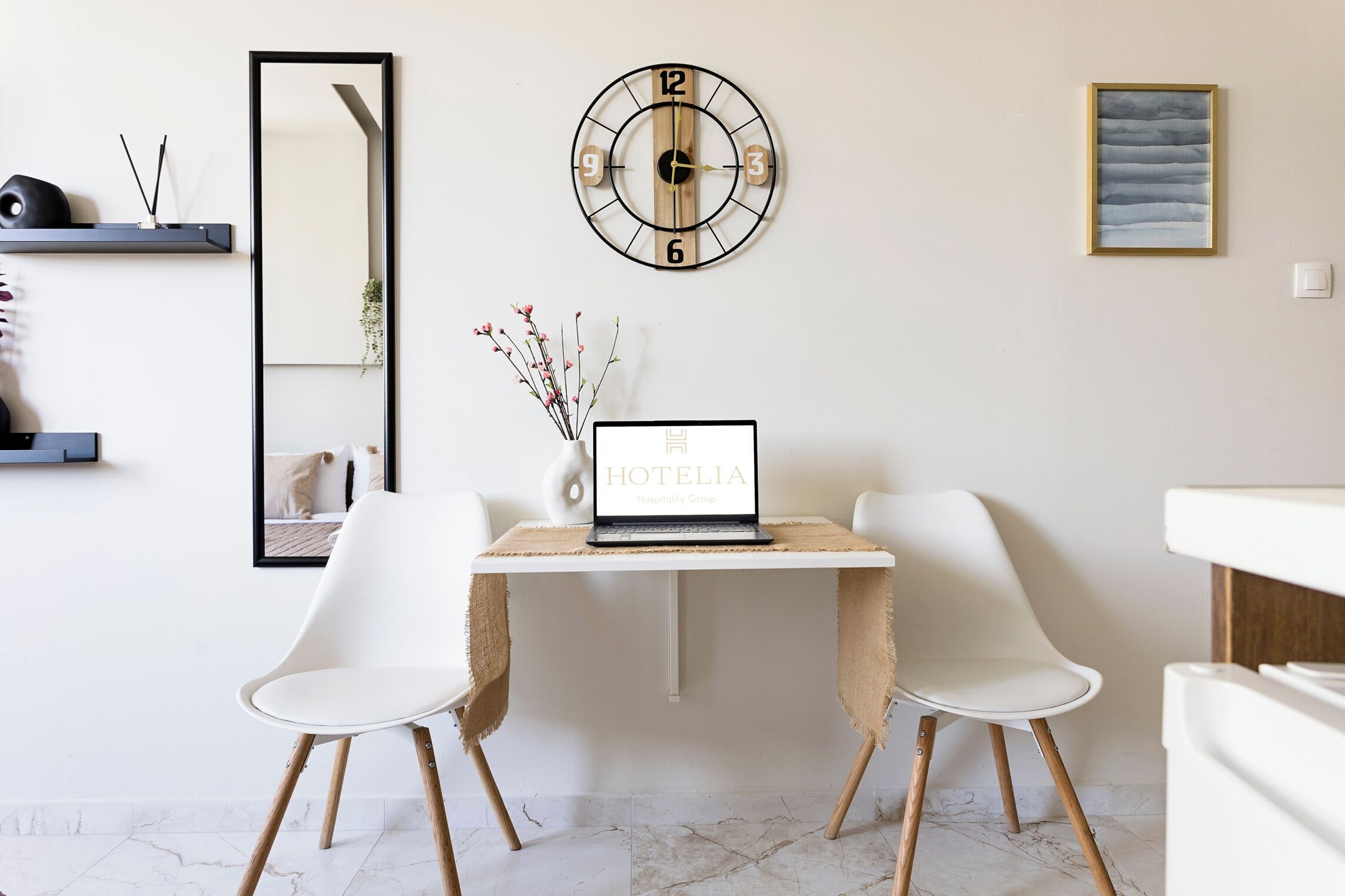 A modern hotel room with a wooden desk, two white chairs, a round wall clock, a framed artwork, and a mirror.