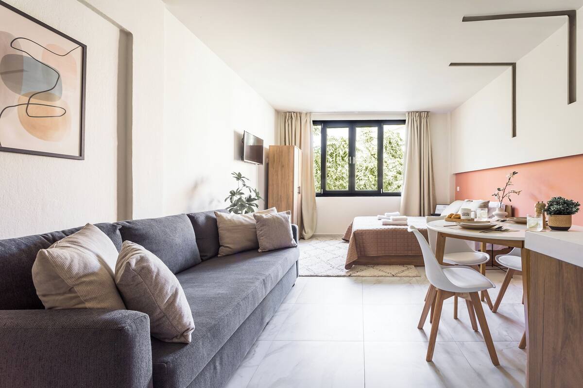 Un salon moderne avec un canapé gris, une table à manger avec des chaises, un téléviseur fixé au mur et une grande fenêtre avec des rideaux.