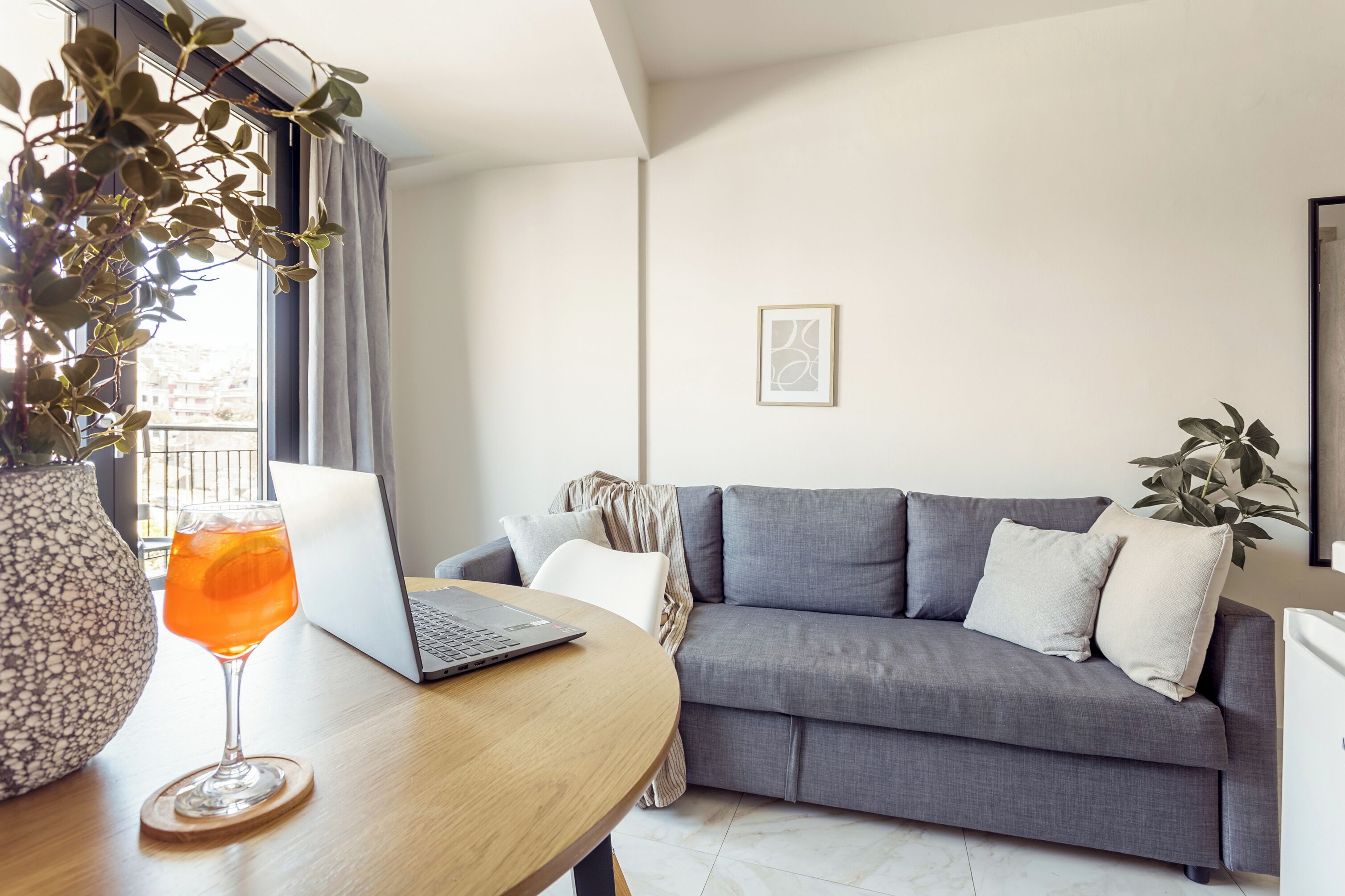 Ein modernes Wohnzimmer mit einem grauen Sofa, einem Holztisch mit einem Laptop und einem Glas Orangensaft, sowie einer Topfpflanze.