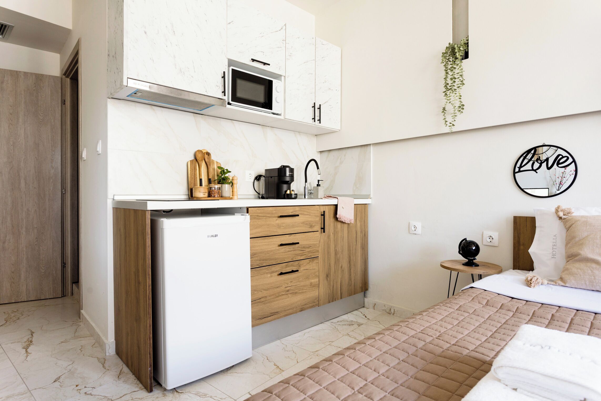 A compact hotel room with a kitchenette, a bed with a beige quilt, and a wall decoration with the word "love".