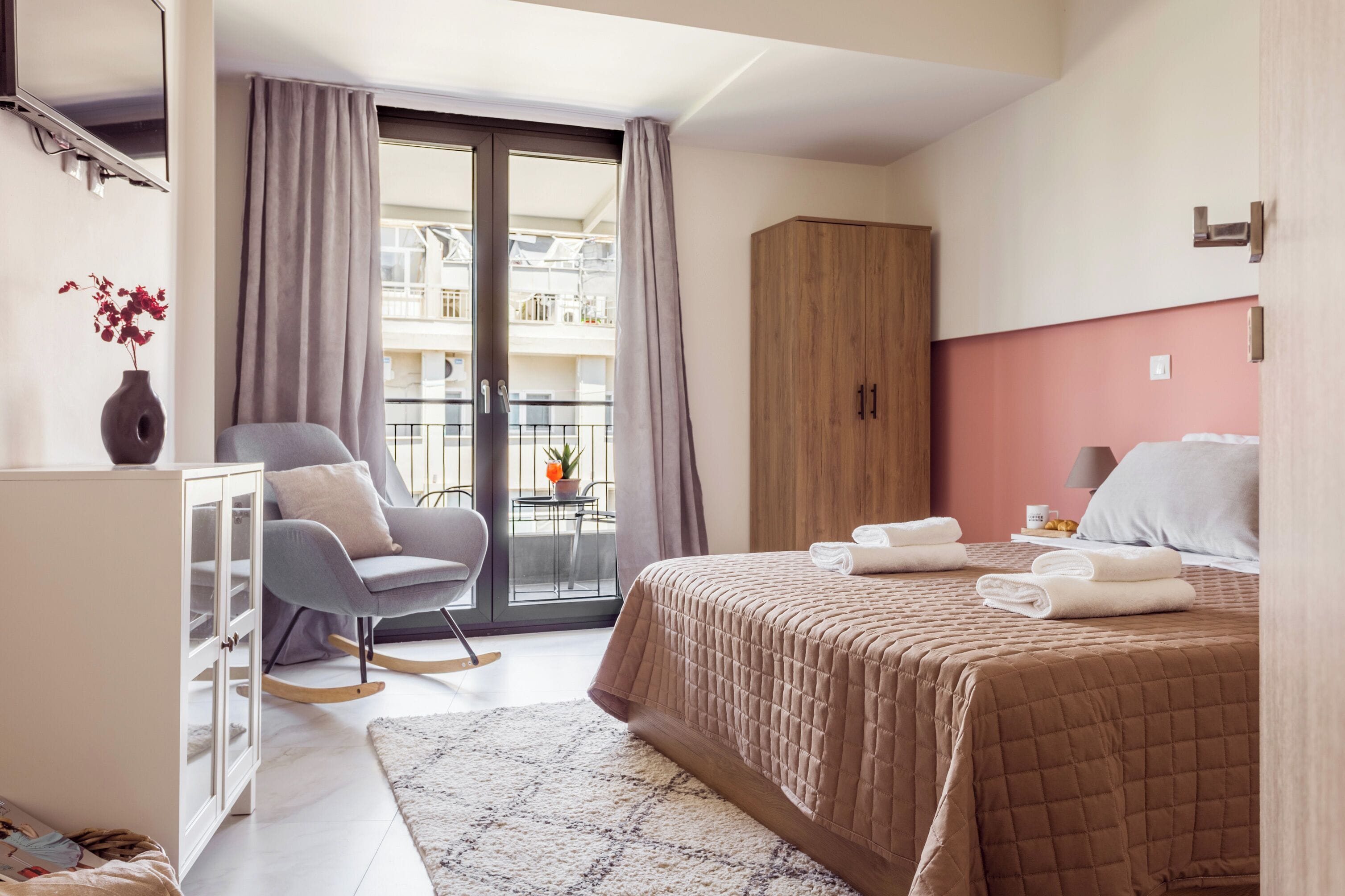 Ein Hotelzimmer mit Bett, Schaukelstuhl, Kleiderschrank, einem kleinen Tisch mit Pflanze und einem Balkon mit Geländer.