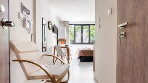 A modern hotel room with a wooden door, a bed with a beige bedspread, a dining area with a table and chairs, and a large window with a view of greenery.