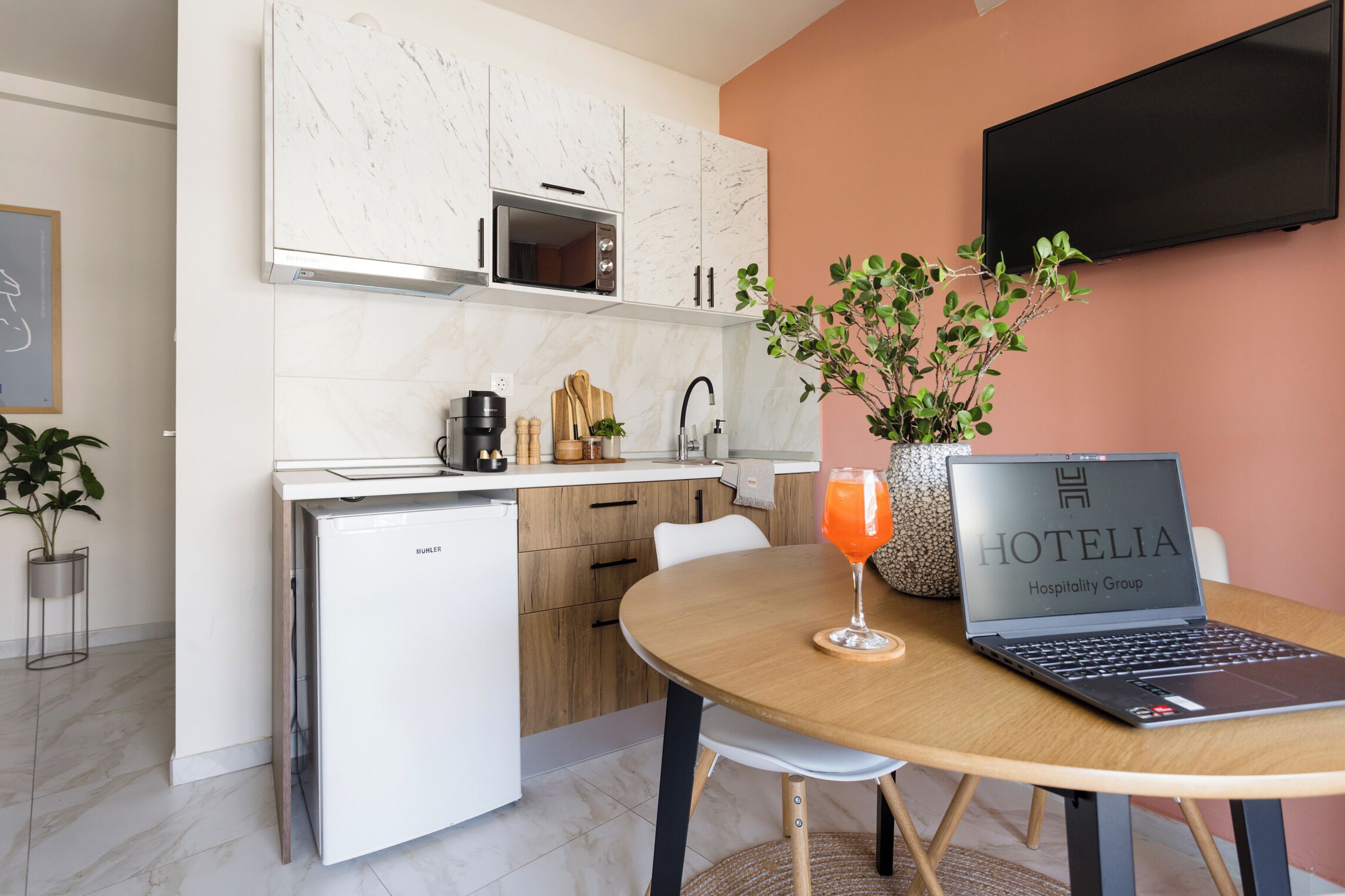 Een compacte keuken met een magnetron, koffiezetapparaat en een kleine koelkast. Er staat een eettafel met een laptop en een glas sinaasappelsap.