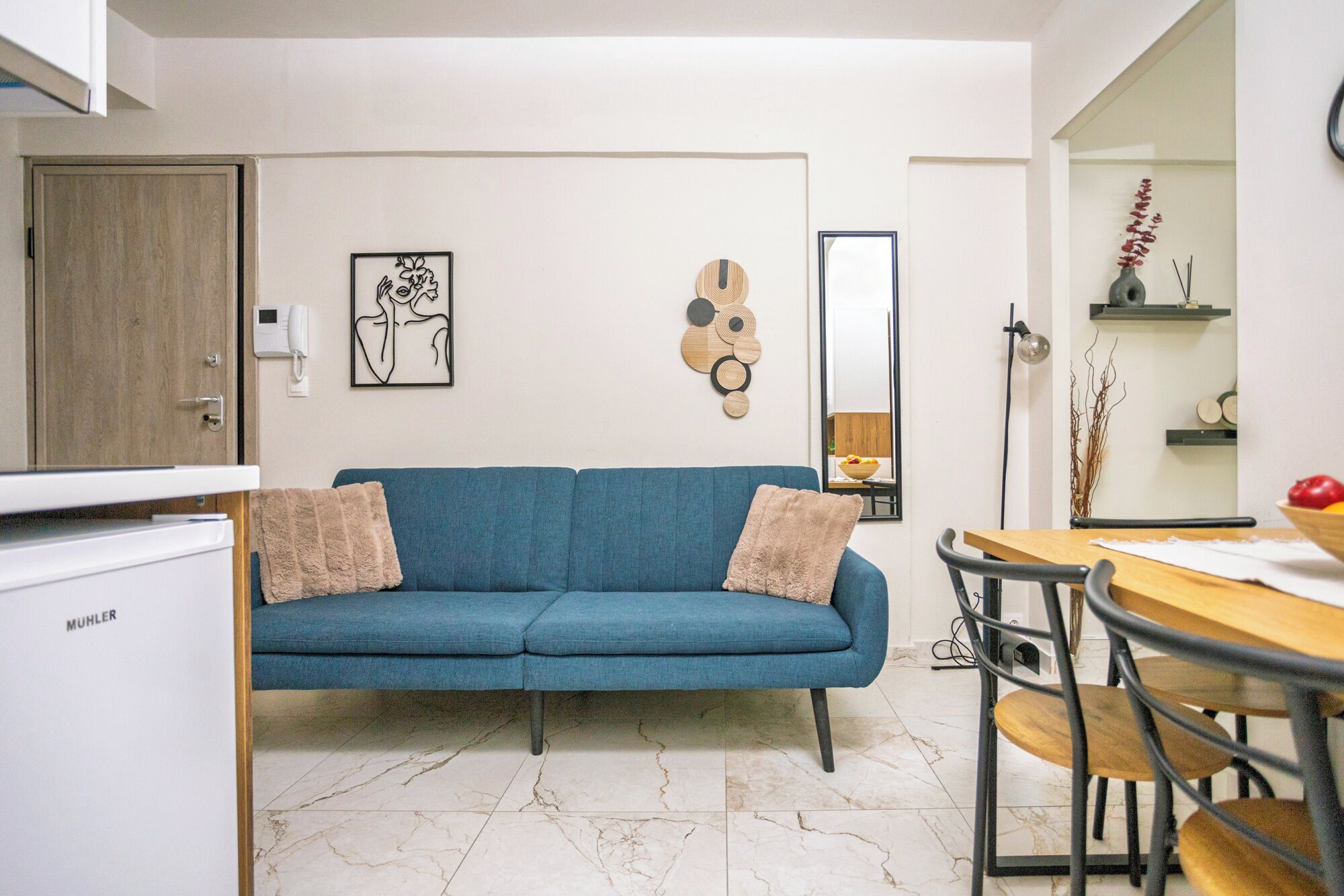 A modern living room with a blue sofa, a dining table with chairs, and a kitchen area with a refrigerator.
