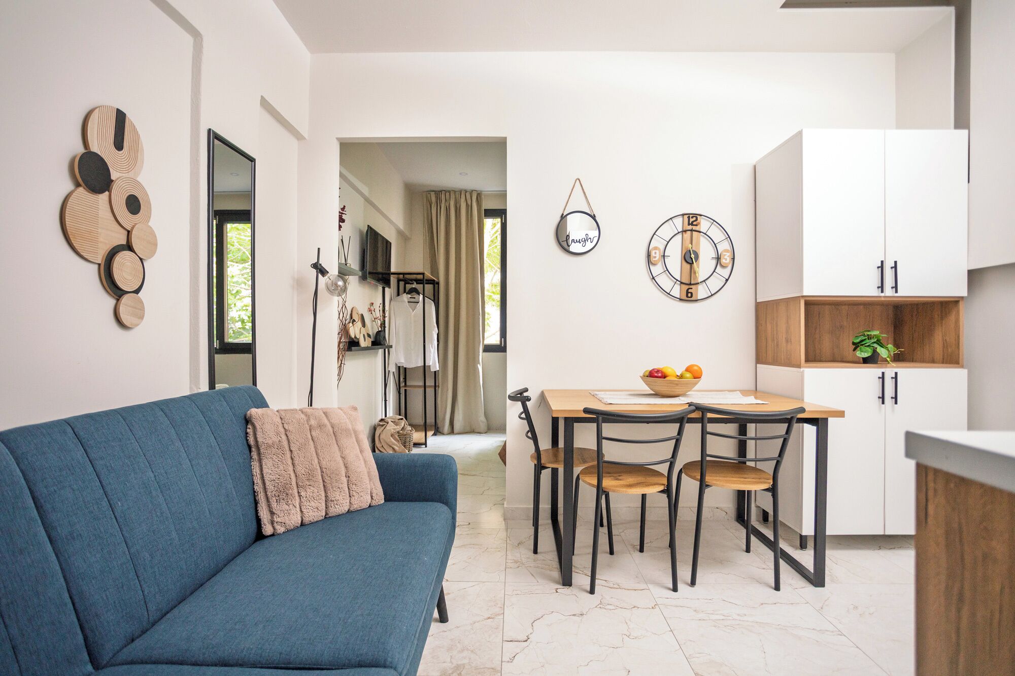 A modern living room with a blue sofa, a dining table with chairs, a kitchen area with cabinets, and a wall clock.
