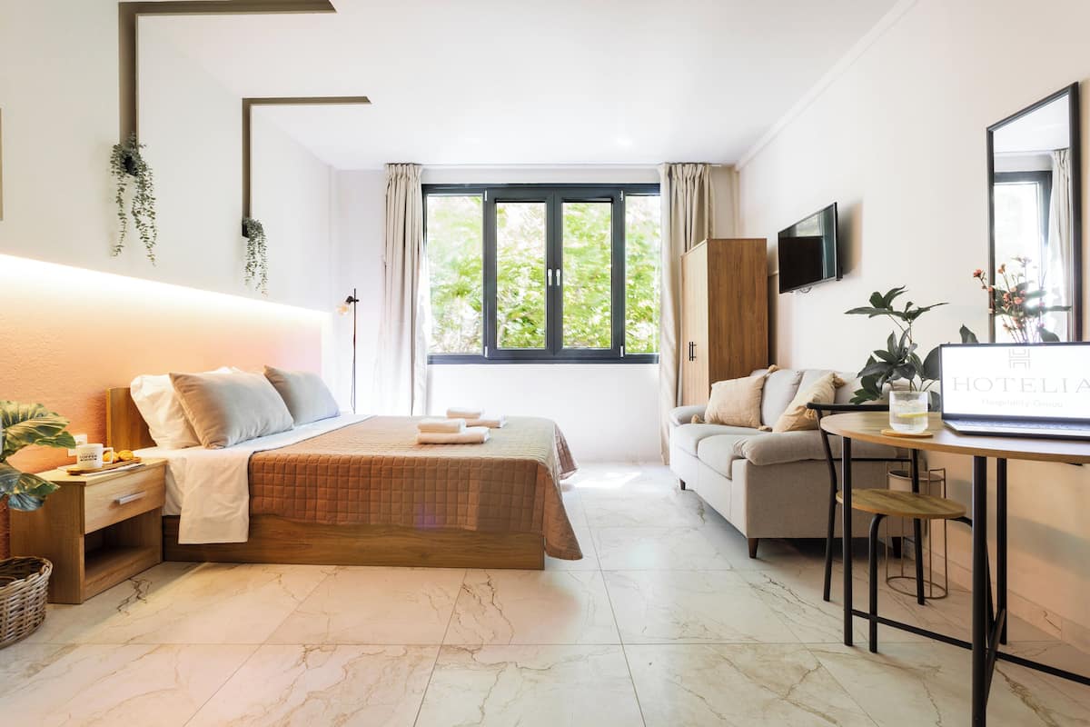 Une chambre d’hôtel moderne équipée d’un lit, d’un canapé, d’un bureau avec un ordinateur portable, d’une télévision et d’une grande fenêtre avec des rideaux.
