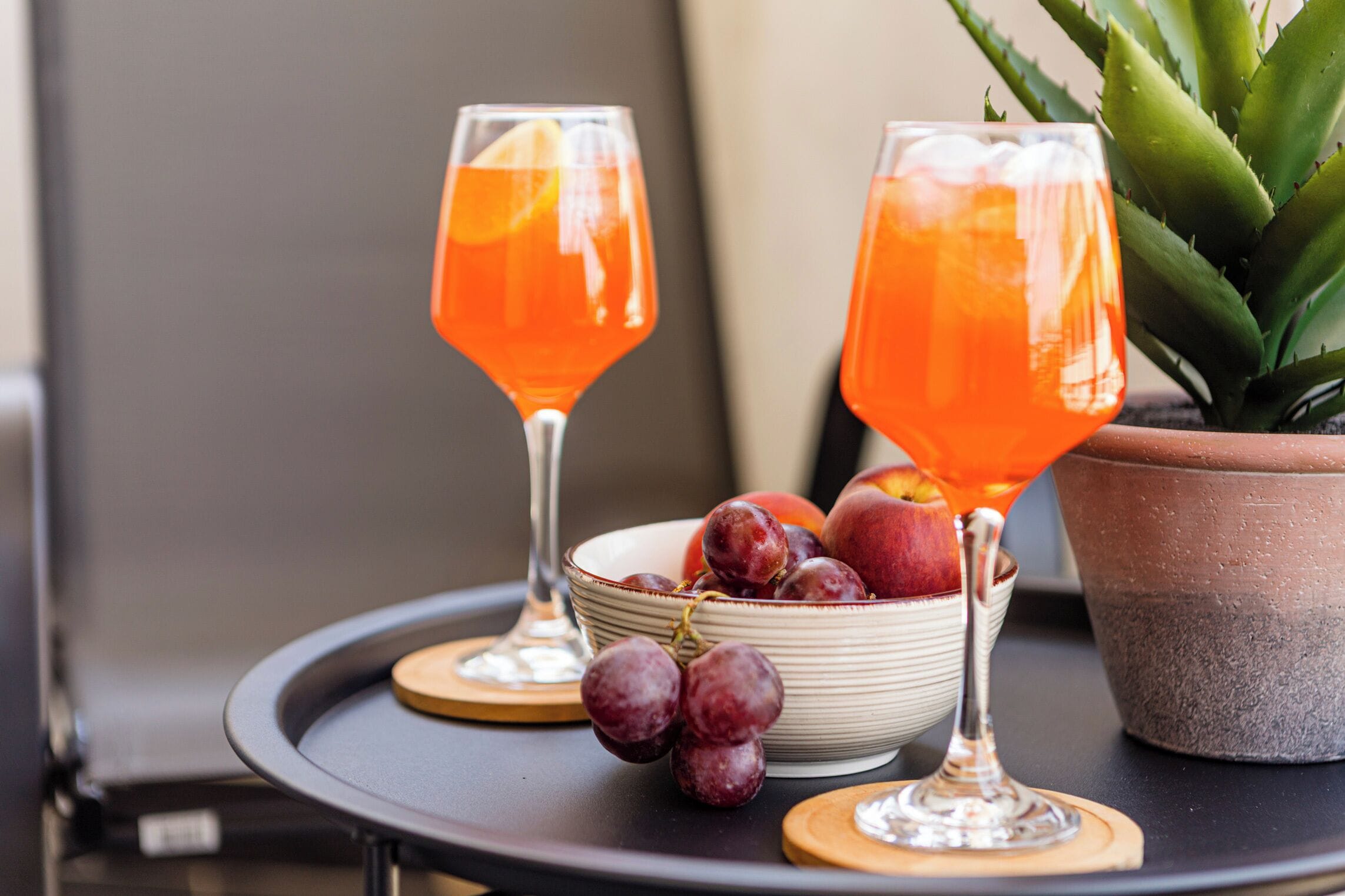 Two glasses of orange beverage on a table with a bowl of fruit and a potted plant.
