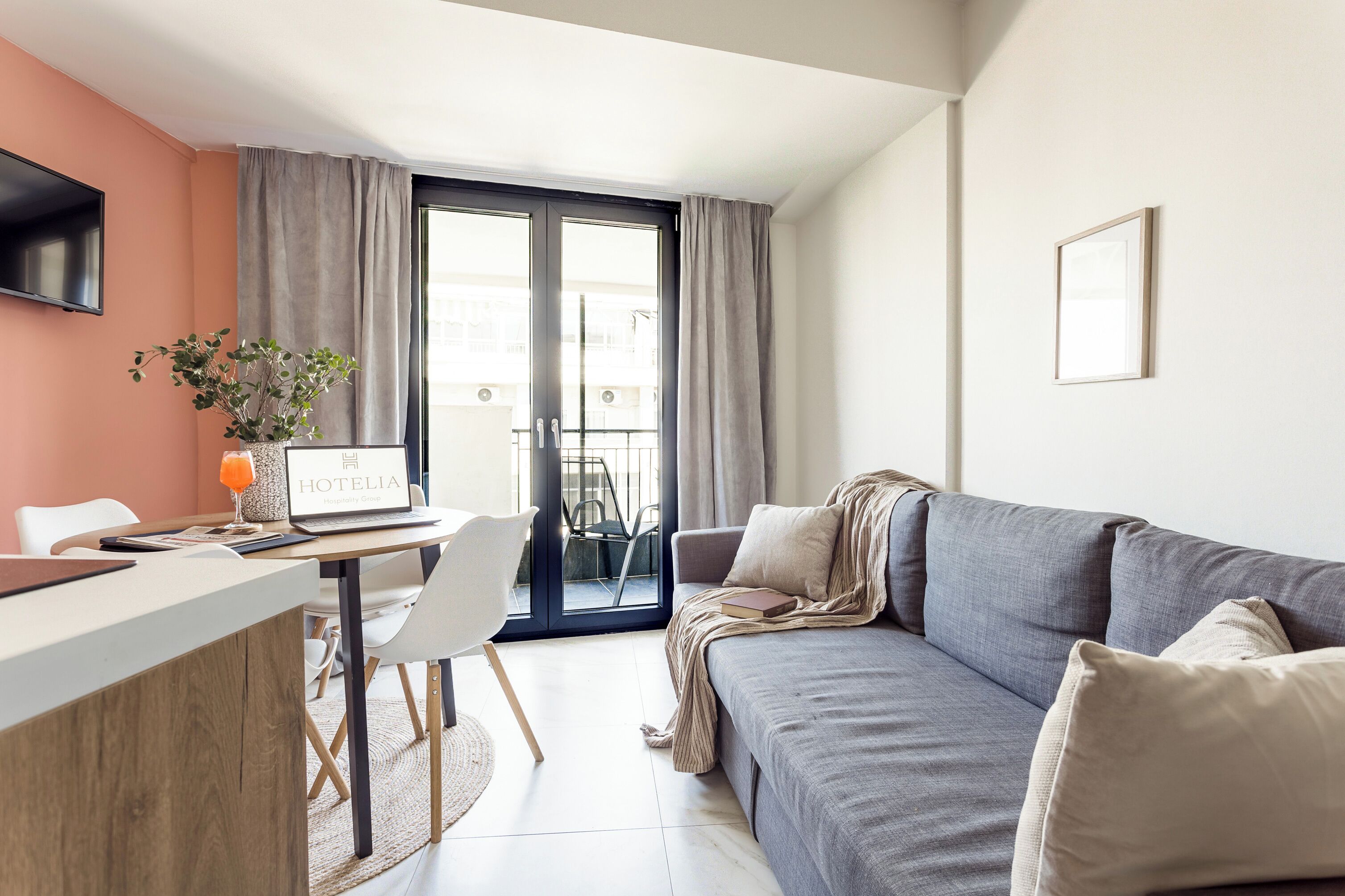 Une chambre d’hôtel moderne avec un canapé gris, un petit bureau avec un ordinateur portable, un téléviseur à écran plat et une plante en pot.