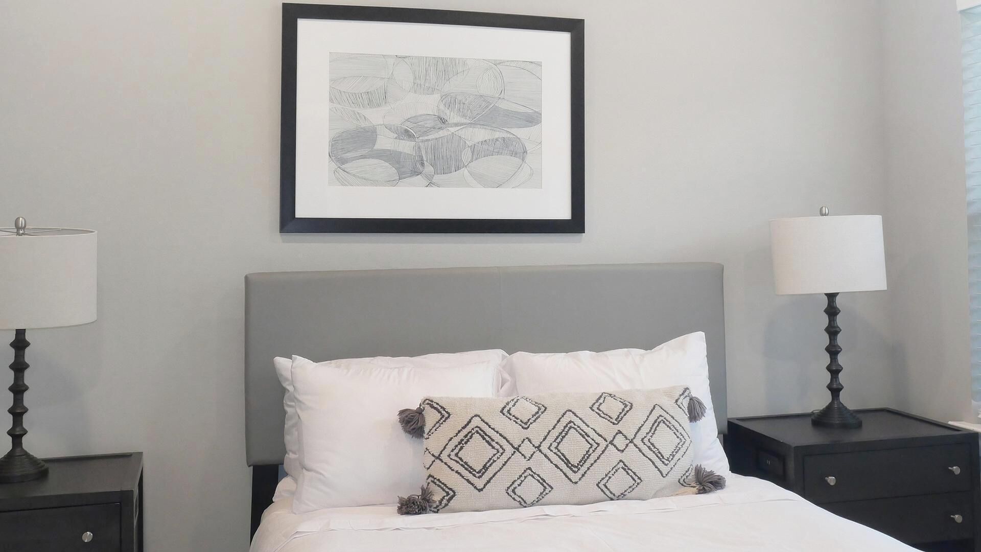 A neatly arranged hotel room with a bed, two bedside tables with lamps, a framed abstract artwork on the wall, and a patterned throw pillow.