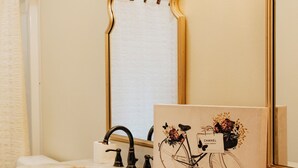 Un baño con encimera de mármol, espejo con marco dorado, toallero y una obra de arte enmarcada de una bicicleta.