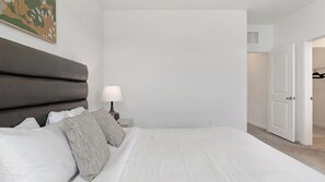 Un dormitorio con una cama grande, una mesita de noche, una lámpara y una puerta que da a otra habitación.