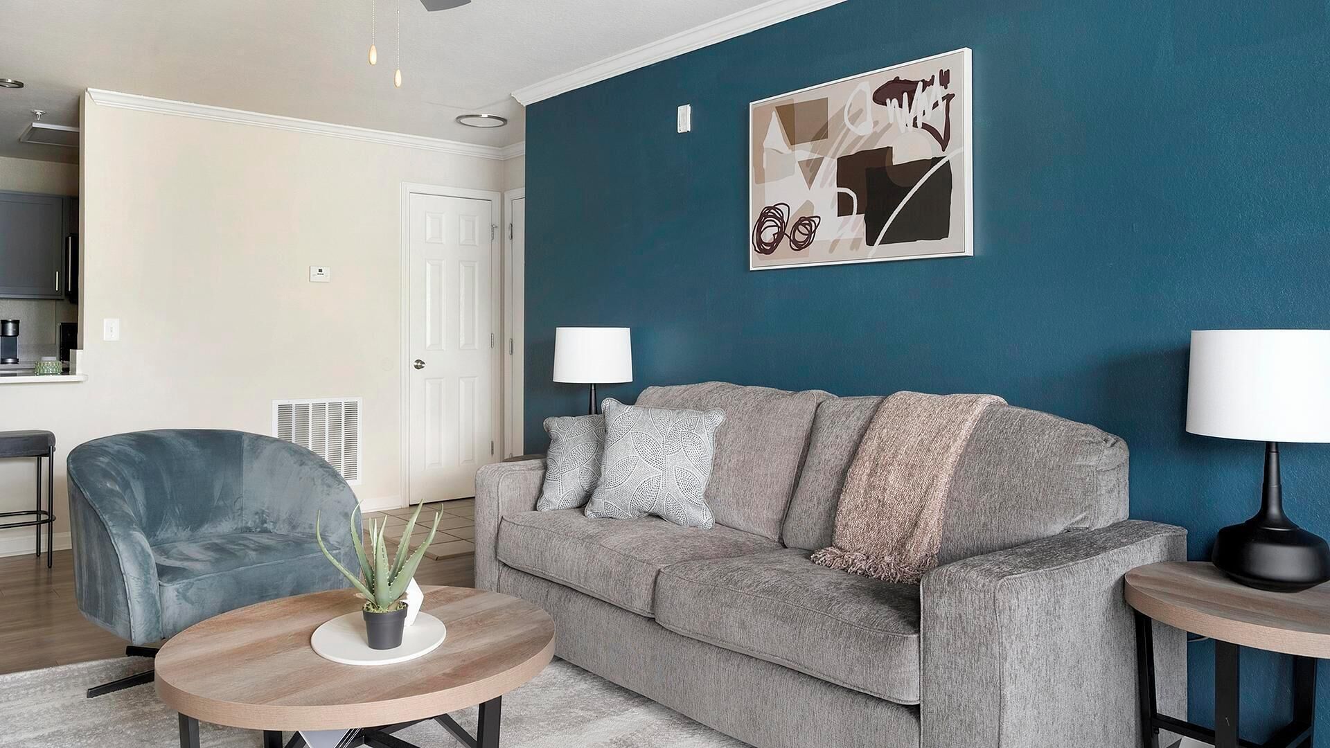 Una sala de estar con un sofá gris, un sillón azul, una mesa de centro redonda de madera y una planta en maceta. Hay un cuadro en la pared y una lámpara sobre una mesita auxiliar.