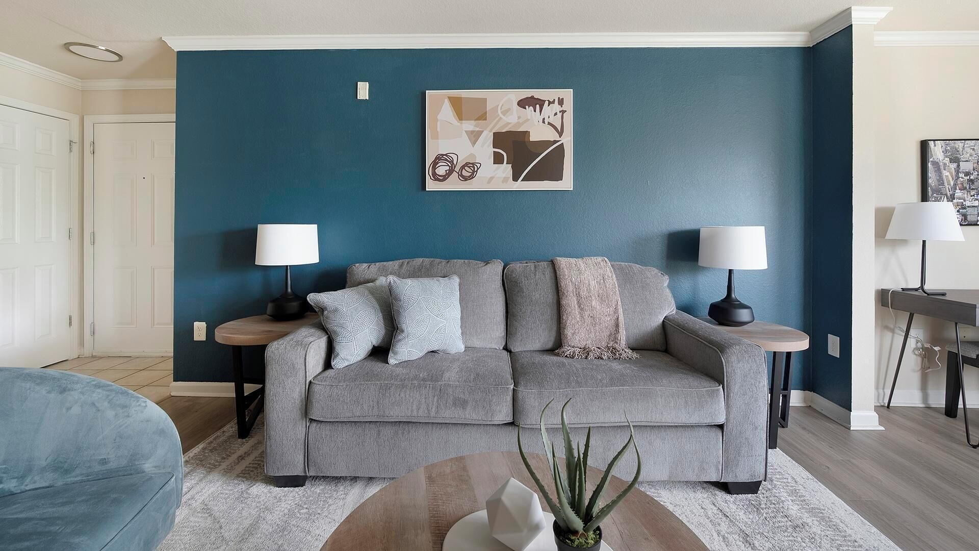 Un salon avec un canapé gris, deux tables d’appoint avec des lampes, un mur d’accent bleu et une œuvre d’art abstraite encadrée.