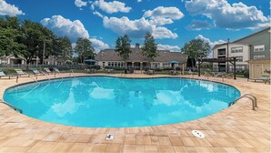 A rectangular swimming pool with a tiled deck, surrounded by lounge chairs and umbrellas, with a clear blue sky and clouds in the background. - Landing | Amazing 2BD, Gym, Game Room (Murfreesboro)