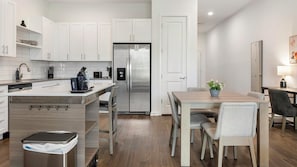 Una cocina moderna con armarios blancos, un refrigerador de acero inoxidable, un lavavajillas integrado y un comedor con mesa y sillas de madera.