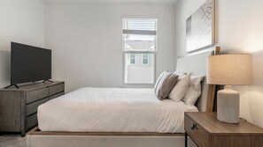 Un dormitorio con cama, televisor sobre un aparador, ventana con persianas y lámpara en la mesita de noche.