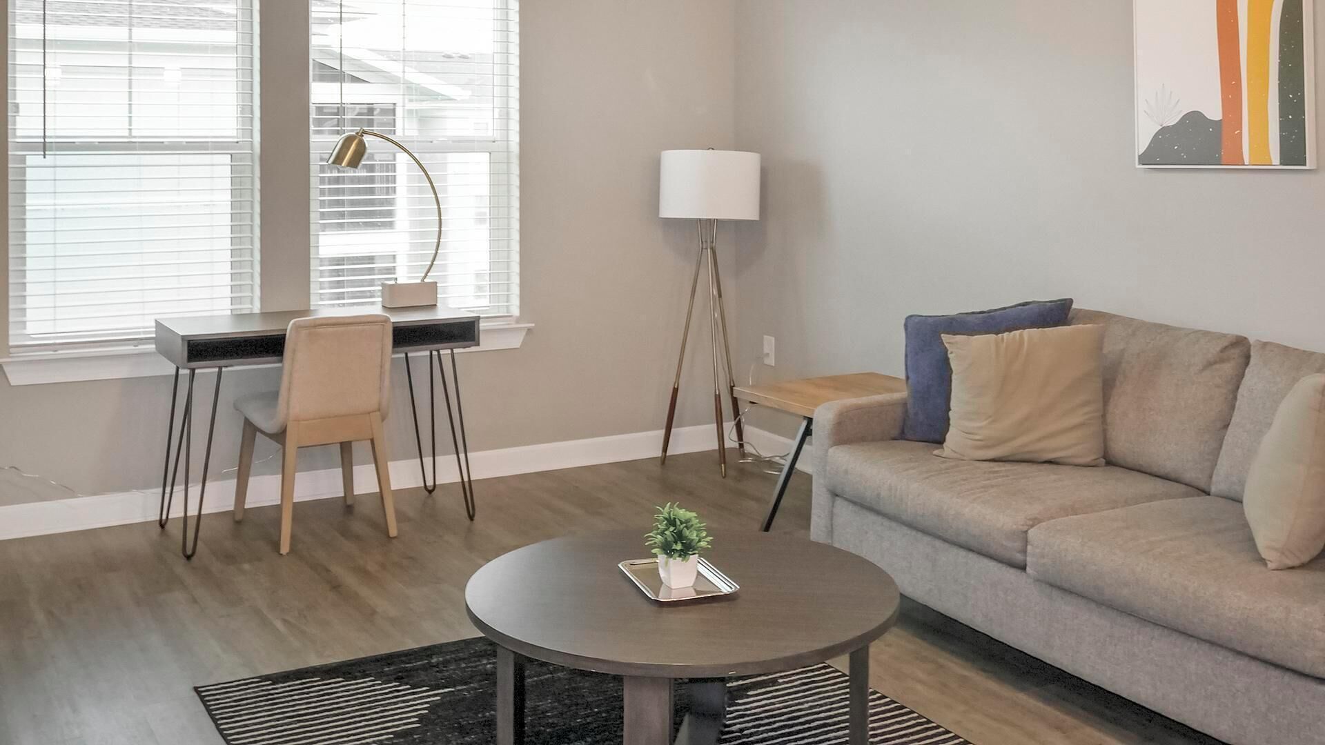 Ein modernes Wohnzimmer mit einer Couch, einem Couchtisch, einem Schreibtisch mit Stuhl, einer Stehlampe und einem Gemälde an der Wand.