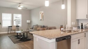 A modern kitchen with granite countertops, a dishwasher, and a coffee maker. There is a dining area with a table and chairs, and a living room with a sofa and a lamp. - Landing | Stylish 1BD, Yoga Studio, Pool (Cape Coral)
