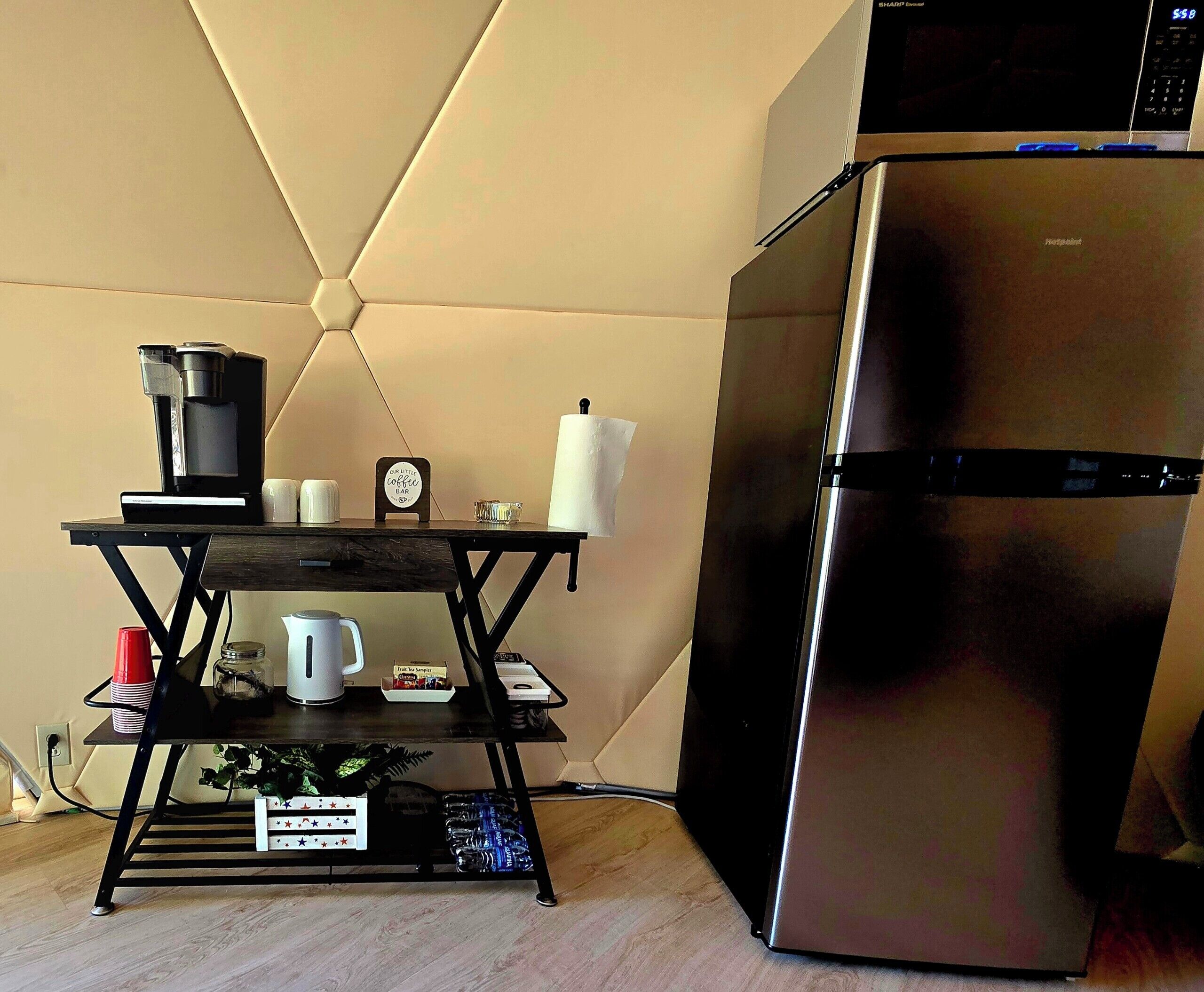 A kitchen with a stainless steel refrigerator, a coffee machine, a kettle, and a small shelf with cups and a sign.