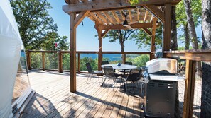 Een houten terras met een pergola, buitenbarbecue en een eethoek met stoelen en een tafel.