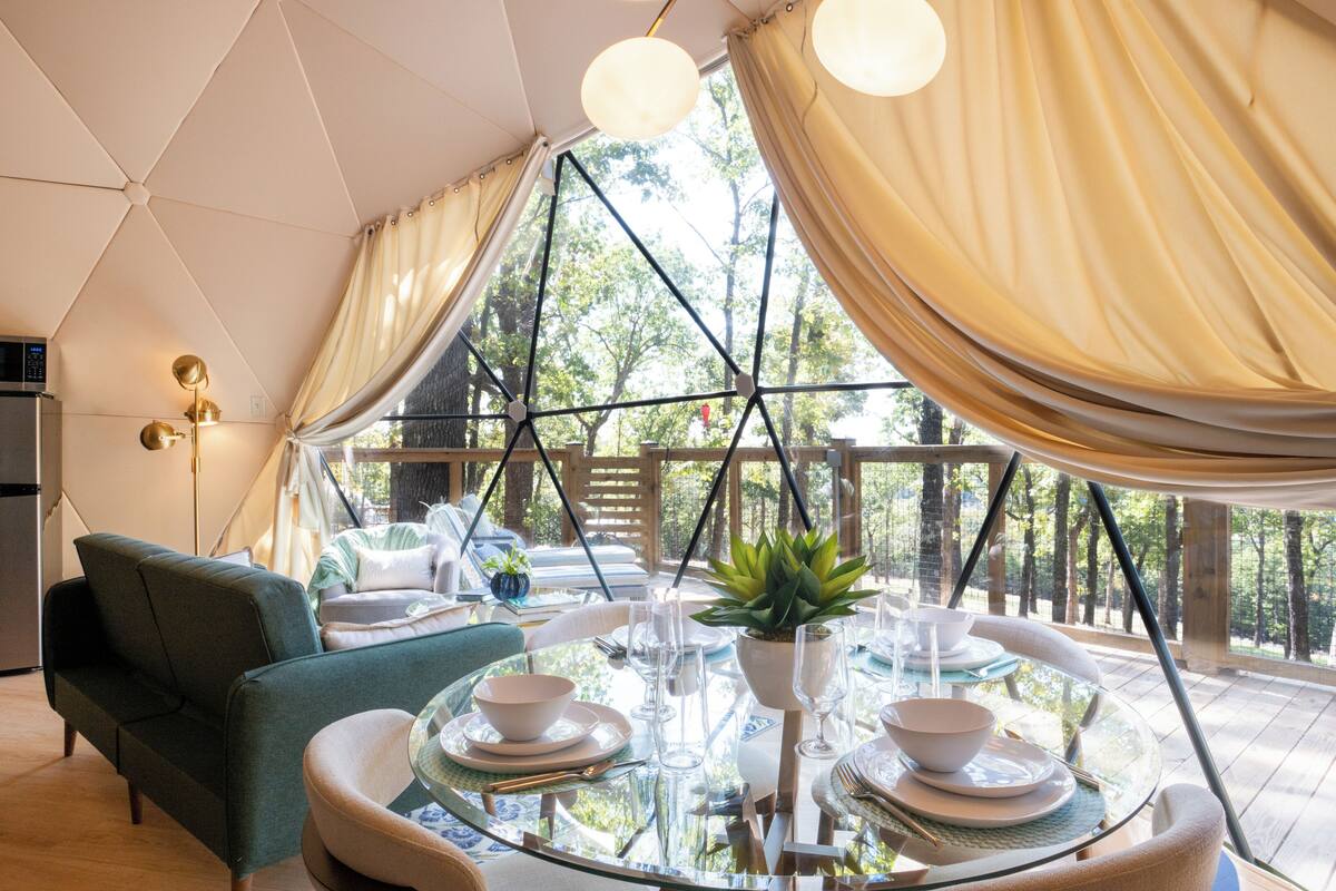 A dining area with a glass table set for four, surrounded by a wooden deck with a view of trees.