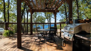 Een overdekt buitengebied met een barbecue, houten terras en uitzicht op het water.