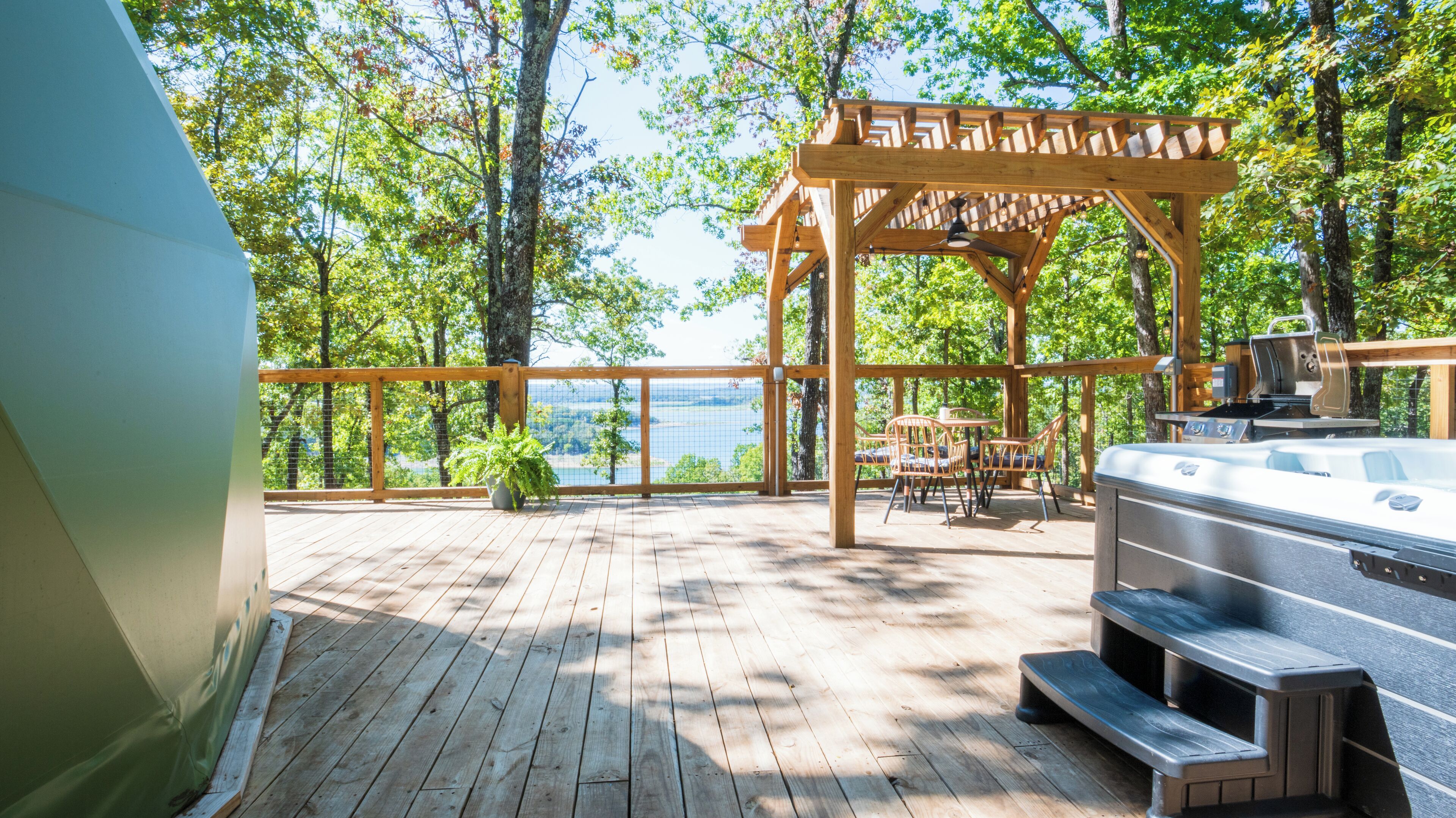 Un terrazzo in legno con una vasca idromassaggio, mobili da esterno e un pergolato con vista su un lago.