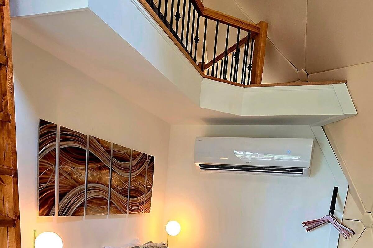 A bedroom with a bed, bedside lamps, a wall-mounted air conditioner, a ceiling fan, and a wooden staircase.