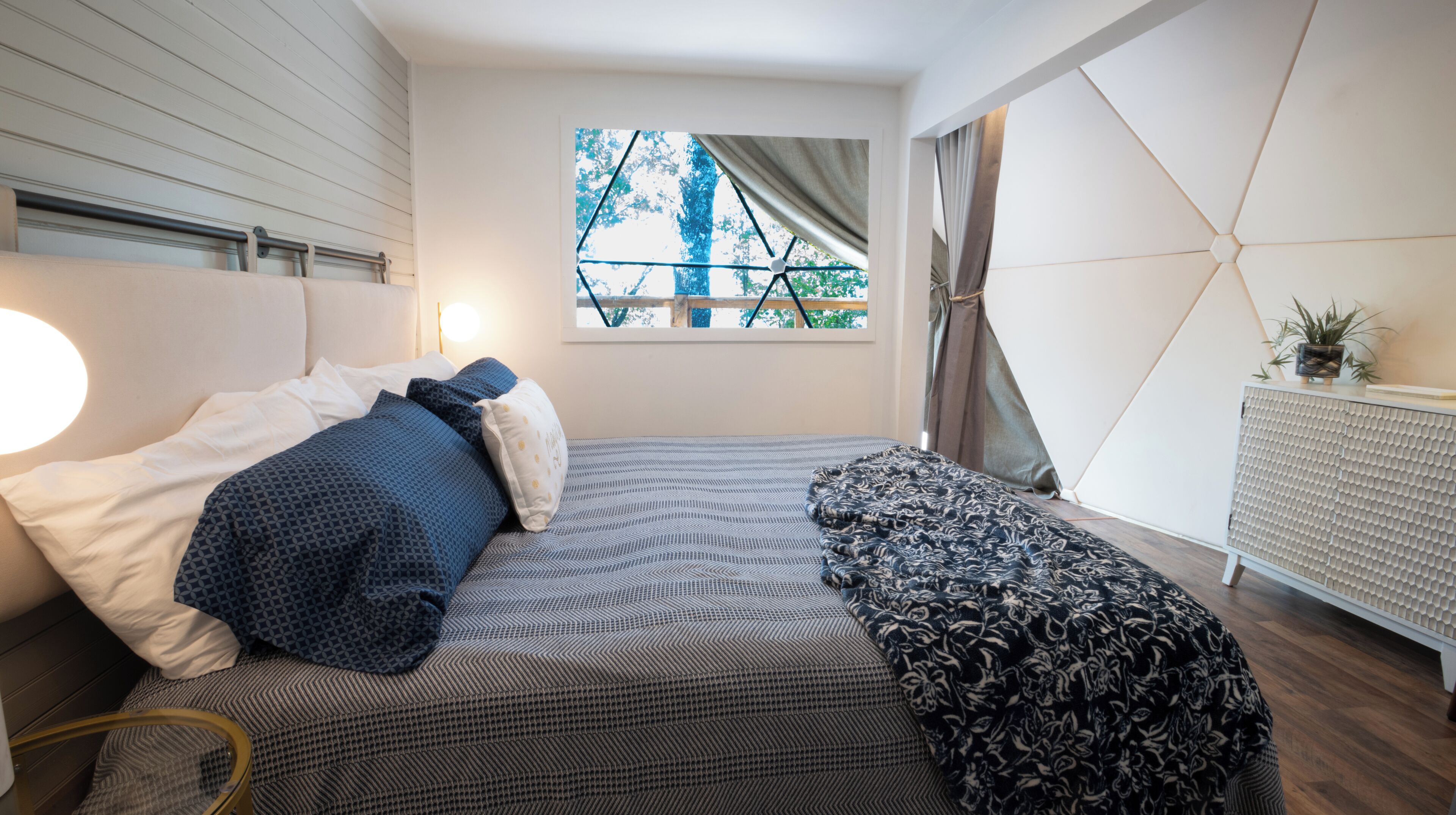 A modern bedroom with a large bed, a window with a view of trees, a radiator, and a bedside table with a plant.