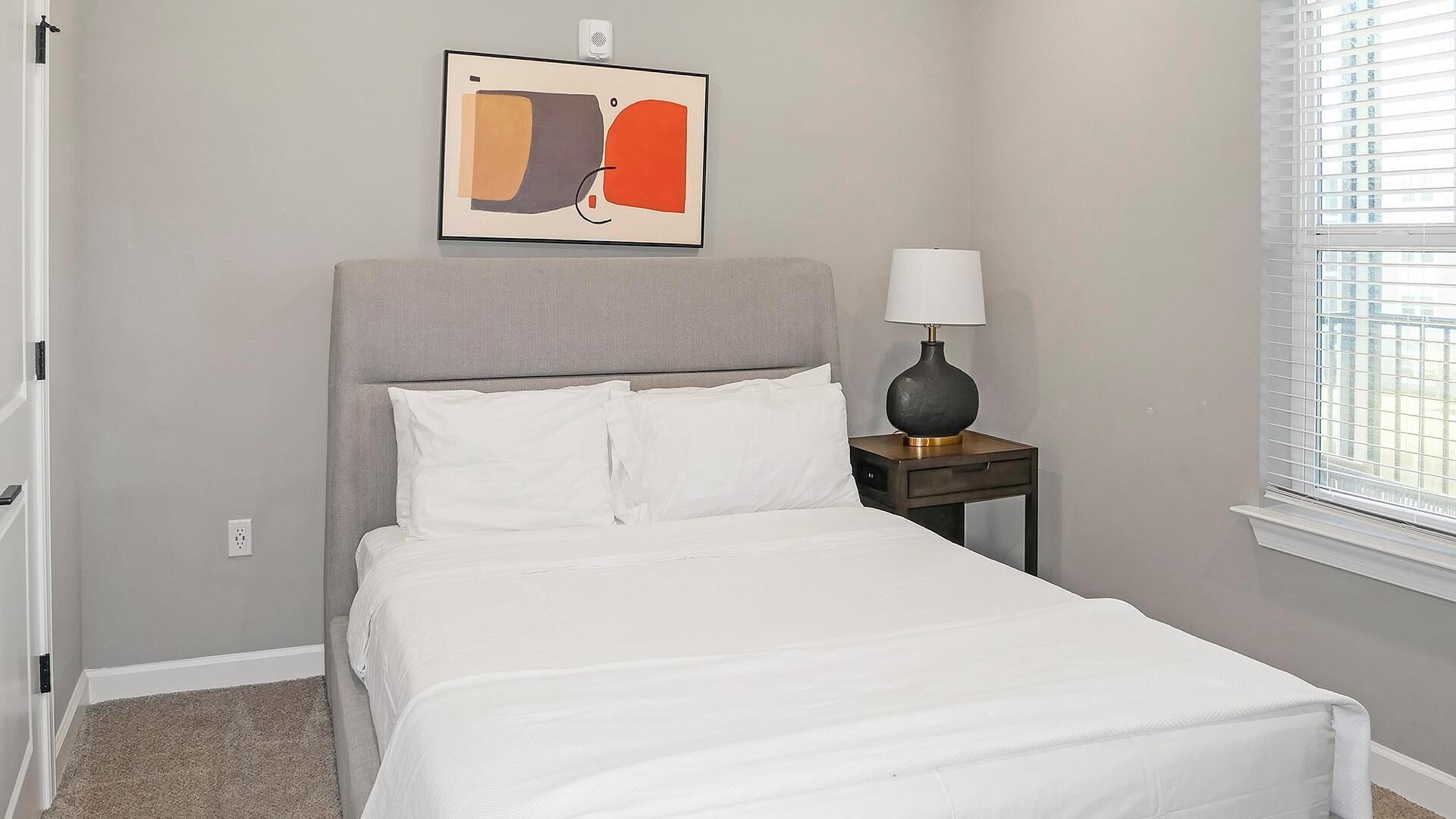 A bedroom with a bed, a nightstand with a lamp, a painting on the wall, and a window with blinds.