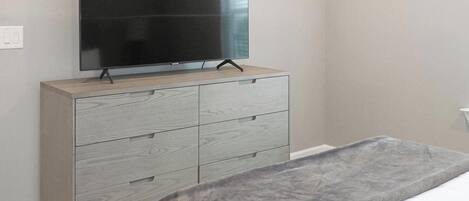 Une chambre à coucher avec une commode en bois, un téléviseur à écran plat fixé au-dessus, et un lit recouvert d’une couverture grise.