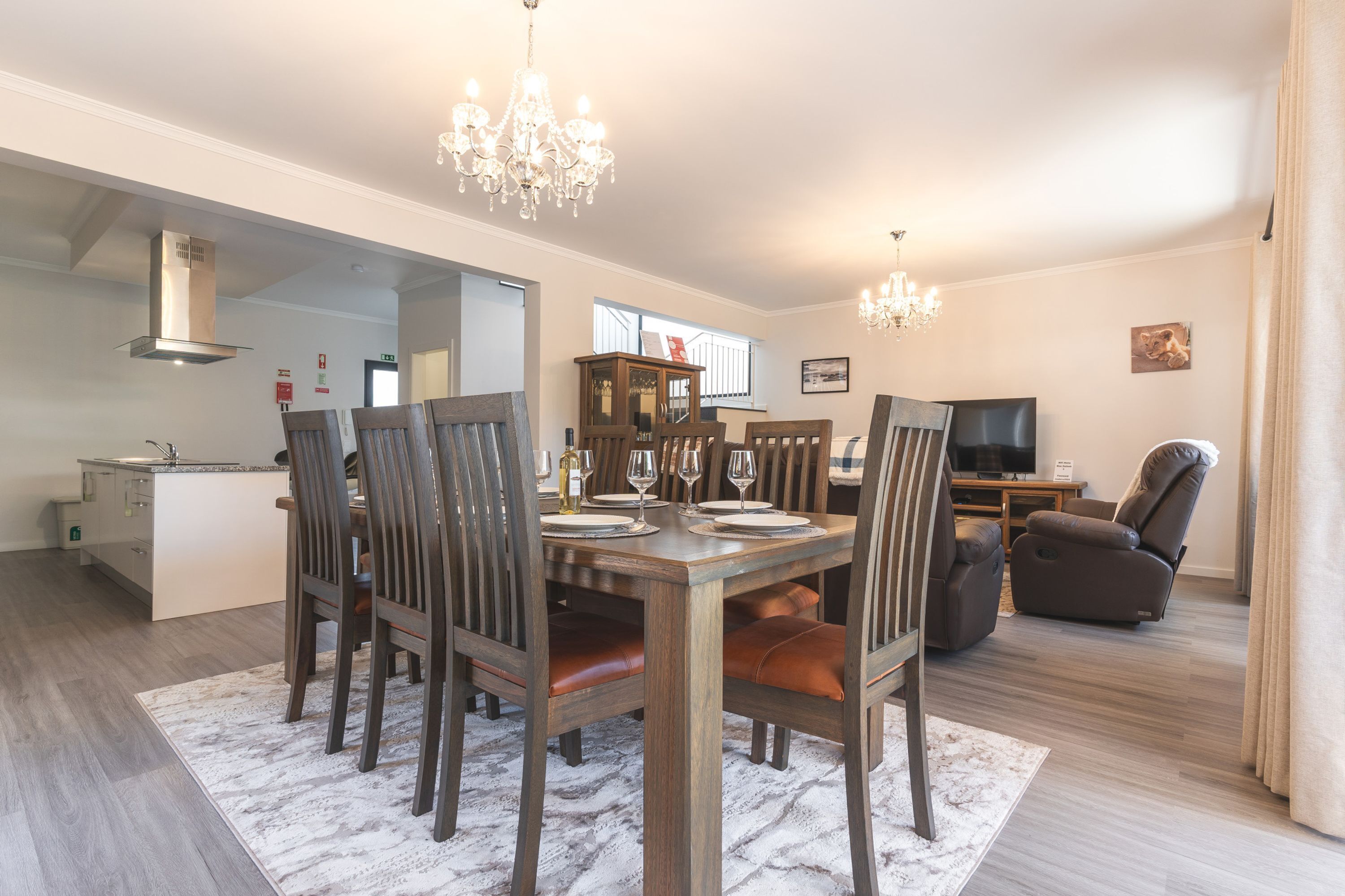 A dining area with a wooden table set for six, surrounded by chairs with leather cushions. A chandelier hangs above, and a television is visible in the background.
