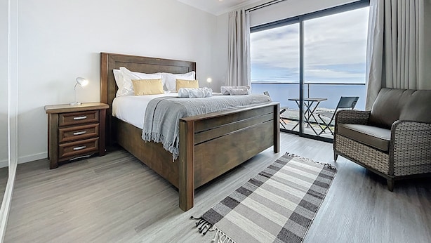 A modern bedroom with a wooden bed, bedside tables, a chair, and a large window offering a view of the sea. - Blue Outlook Retreat by Atlantic Holiday (Santa Cruz)